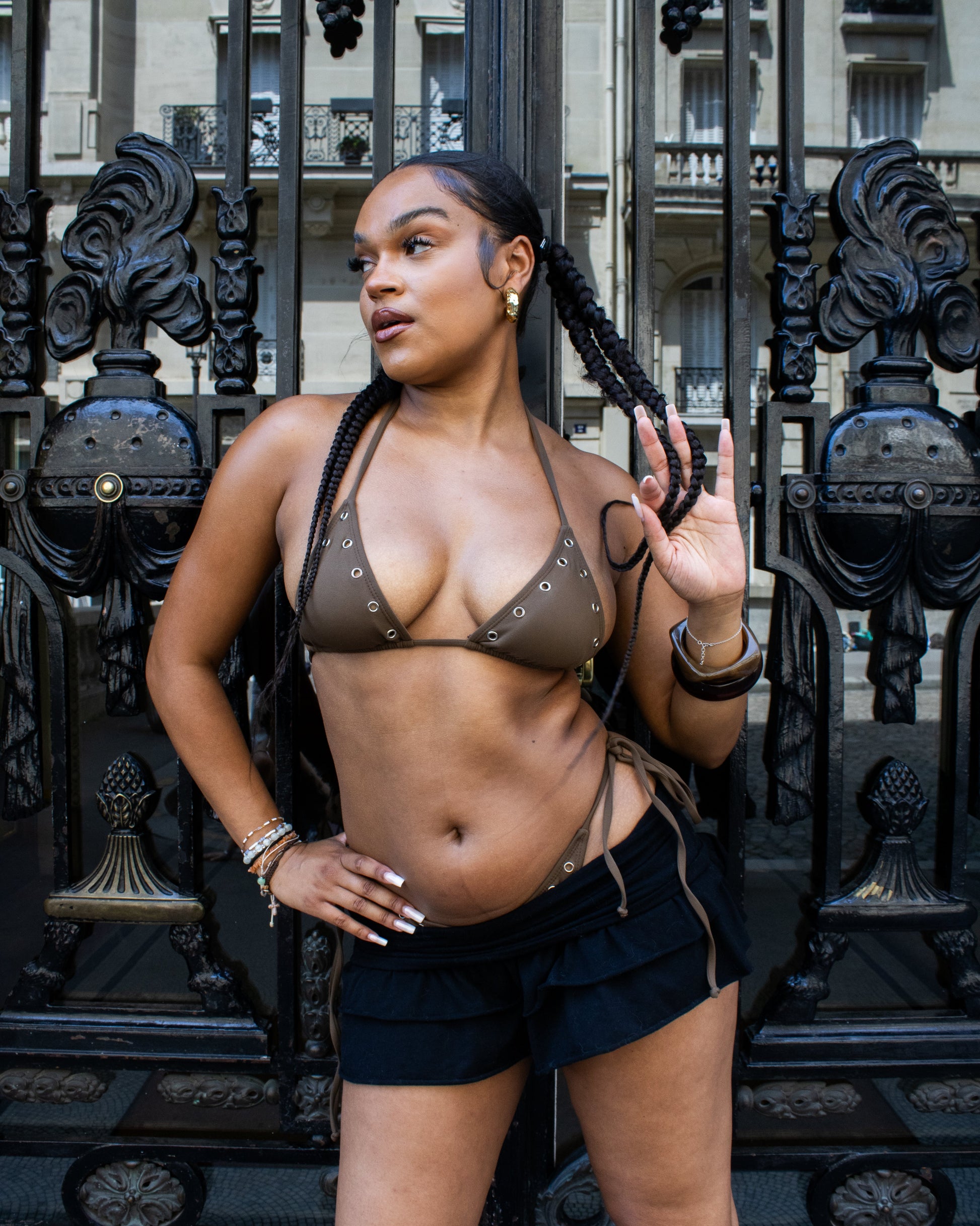 A woman posing in front of paris gates, wearing a brown halter bikini top with silver eyelet details and black shorts.